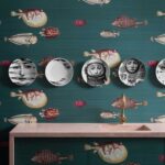 Wand met Fornasetti Acquario-behang met kleurrijke vismotieven en decoratieve borden met surrealistische gezichten boven een roze wastafel.