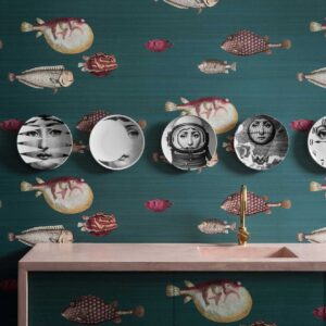 Wand met Fornasetti Acquario-behang met kleurrijke vismotieven en decoratieve borden met surrealistische gezichten boven een roze wastafel.