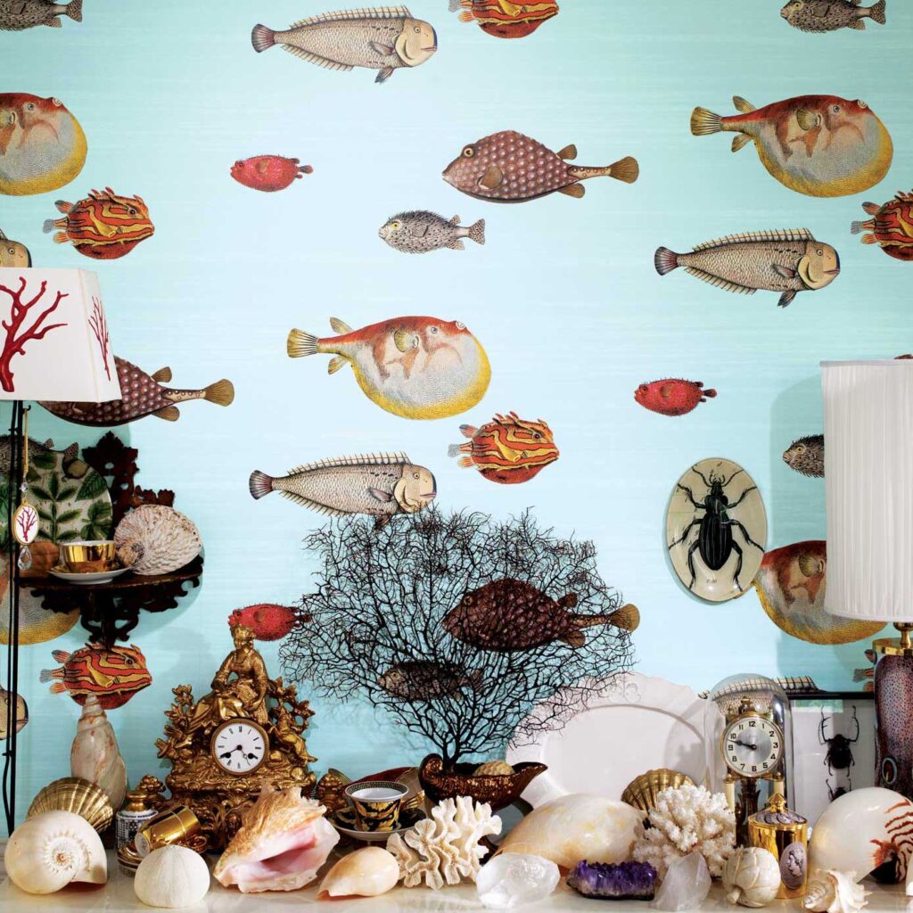 Fornasetti Acquario behang met kleurrijke illustraties van vissen als achtergrond voor een eclectische verzameling schelpen, klokken en decoratieve objecten.