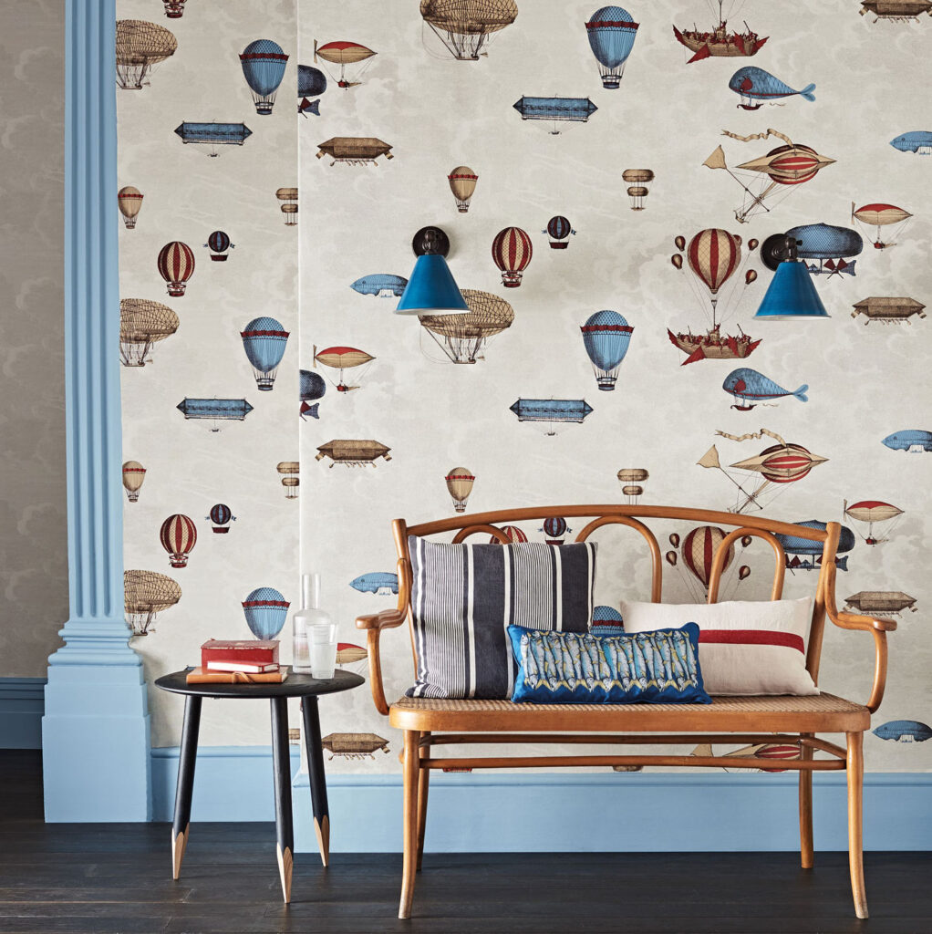 Houten bankje met kussens voor een muur met Fornasetti Macchine Volanti-behang vol fantasierijke luchtballonnen en vliegmachines.