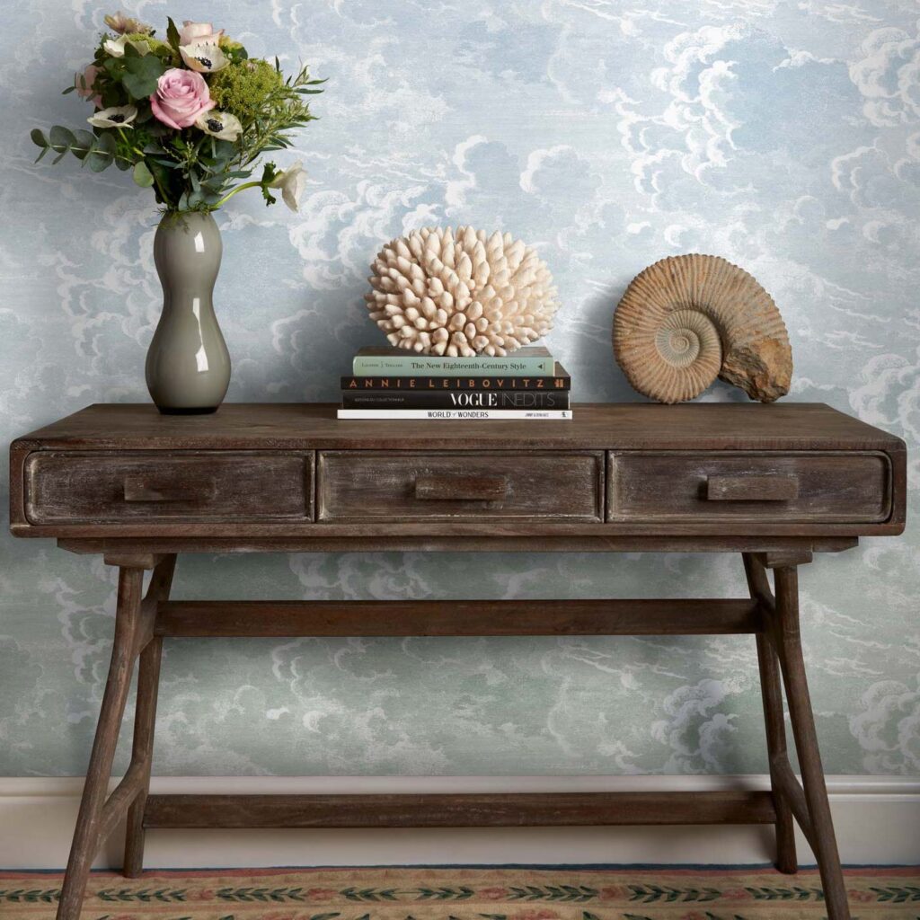 Een houten consoletafel met decoratieve objecten staat tegen Fornasetti-behang Nuvole al Tramonto met een dromerig wolkenpatroon.