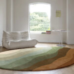 Ronde Limited Edition Allegro Low Honey Pesto vloerkleed met aardetinten in een minimalistisch interieur met een witte lounge stoel en een ronde bijzettafel.