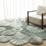 Organisch gevormd Atoll vloerkleed in aqua tinten met een houten fauteuil erop in een lichte, sfeervolle ruimte.
