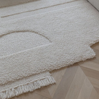 Detail van een limited edition vloerkleed met abstract organisch rechthoekig ontwerp in een zachte, crèmekleurige stof op een houten visgraatvloer.