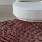 Rond vloerkleed Spiro in auburn bruin met een reliëfpatroon, geplaatst voor een ronde, lichtkleurige bank.