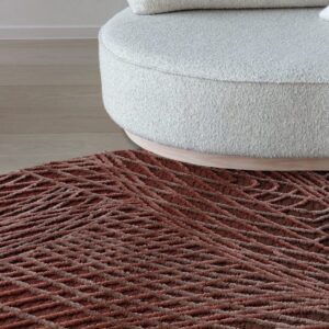 Rond vloerkleed Spiro in auburn bruin met een reliëfpatroon, geplaatst voor een ronde, lichtkleurige bank.