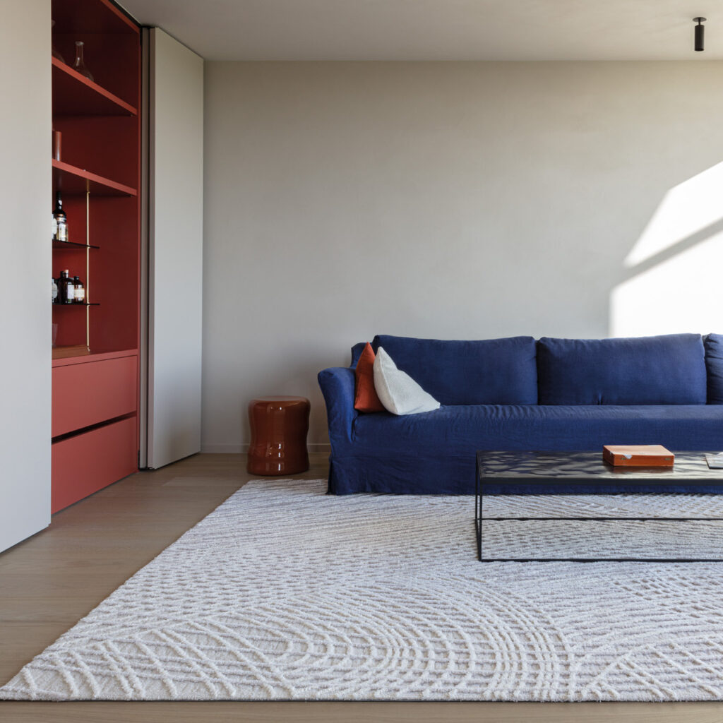 Moderne woonkamer met een Limited Edition vloerkleed Spiro in de kleur Vanilla Fog, een donkerblauwe bank en een glazen salontafel.