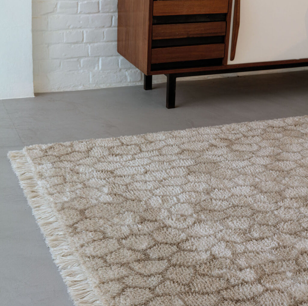 Beige Limited Edition vloerkleed Cosmic Mongolia Papyrus met franjes op een betonnen vloer naast een houten dressoir.