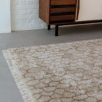 Beige Limited Edition vloerkleed Cosmic Mongolia Papyrus met franjes op een betonnen vloer naast een houten dressoir.