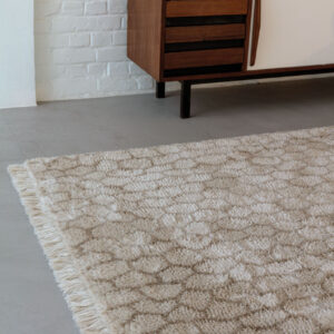 Beige Limited Edition vloerkleed Cosmic Mongolia Papyrus met franjes op een betonnen vloer naast een houten dressoir.