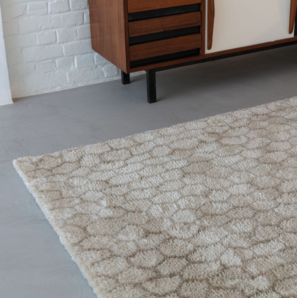 Beige Limited Edition vloerkleed Cosmic Mongolia Papyrus met reliëfpatroon op een grijze betonvloer, naast een houten dressoir.