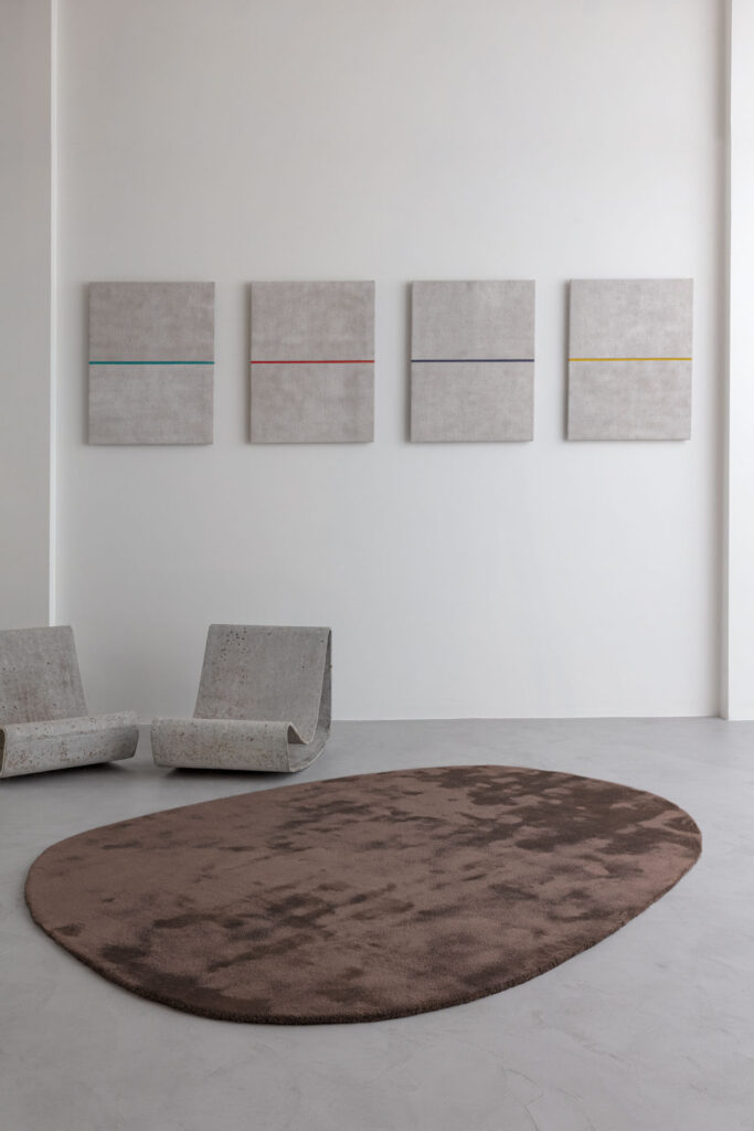 Limited edition vloerkleed Astral Cinder in een minimalistische ruimte met betonnen stoelen en abstracte wandkunst.