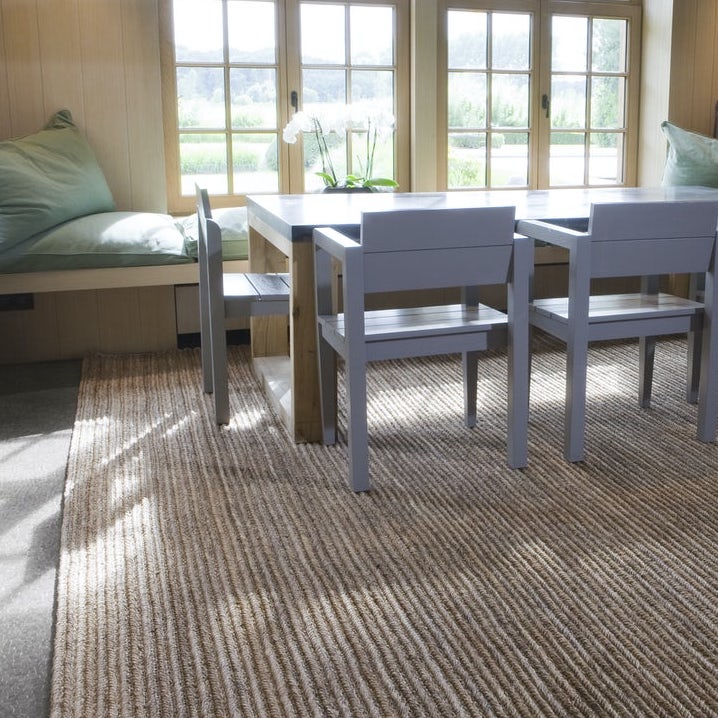 Lichte eetkamer met een limited edition vloerkleed van abacavezels onder een houten tafel en stoelen bij een raam.