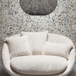 Crèmekleurige ronde loveseat met drie kussens voor een wand met Moooi-behang Armoured Boar-patroon en een zwarte hanglamp erboven.
