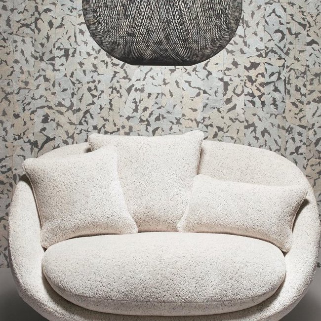 Crèmekleurige ronde loveseat met drie kussens voor een wand met Moooi-behang Armoured Boar-patroon en een zwarte hanglamp erboven.