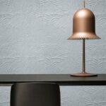 Mat lichtblauw Moooi Dodo Pavone-behang met reliëfpatroon achter een zwarte tafel en een koperen designlamp.