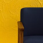 Donkerblauwe stoel met houten armleuning voor een gele muur met reliëfpatroon uit het Moooi-behang Dodo Pavone.