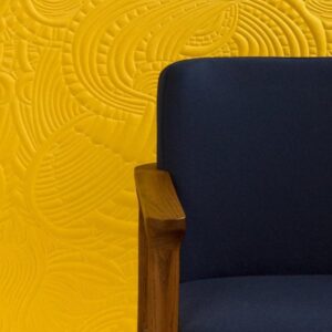 Donkerblauwe stoel met houten armleuning voor een gele muur met reliëfpatroon uit het Moooi-behang Dodo Pavone.