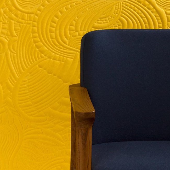 Donkerblauwe stoel met houten armleuning voor een gele muur met reliëfpatroon uit het Moooi-behang Dodo Pavone.