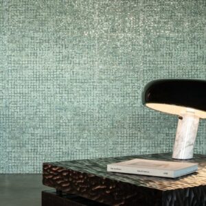Glanzende groenblauwe mozaïektegels uit de Mosaico4-IGG1-2 collectie vormen een elegante wand achter een designlamp en een tafel met boeken.