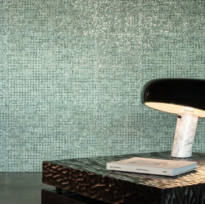Glanzende groenblauwe mozaïektegels uit de Mosaico4-IGG1-2 collectie vormen een elegante wand achter een designlamp en een tafel met boeken.