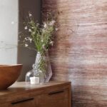 Vaas met veldbloemen op een houten kast naast een houten kom en een glazen stolp met kaars, tegen een gemêleerde roze wand.