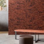 Moderne interieursetting met een rechthoekige wandbekleding in het ontwerp ‘Orrizonte1-3WDD-2’ in rood-bruine tinten en een rood tafelblad op metalen poten.