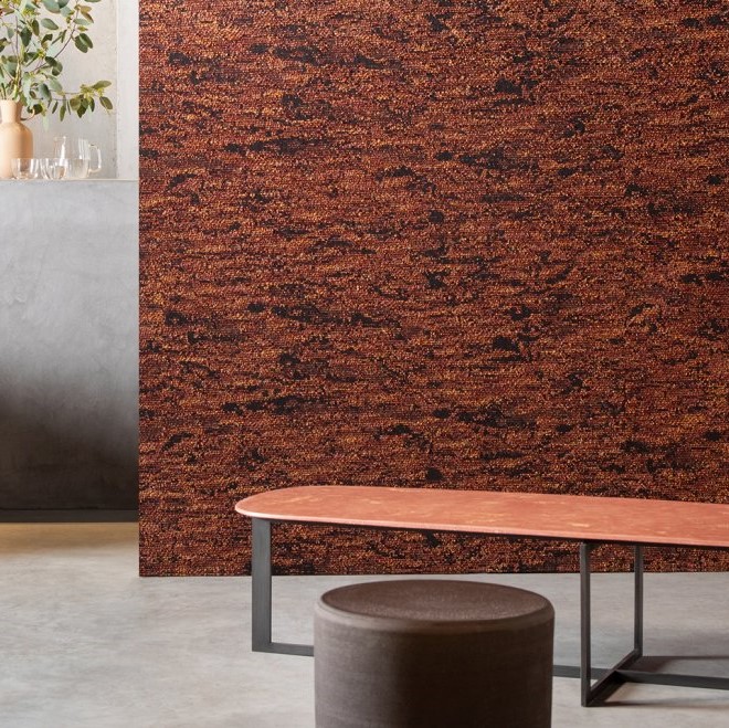 Moderne interieursetting met een rechthoekige wandbekleding in het ontwerp ‘Orrizonte1-3WDD-2’ in rood-bruine tinten en een rood tafelblad op metalen poten.