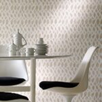 Witte designstoelen en keramiek op een ronde tafel voor een sierlijk beige Perles-Topaze behang met bladpatroon.