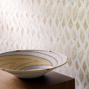 Decoratieve schaal met geweven patroon op houten oppervlak, tegen een wand met Perles Topaze-behang met goud en crème structuurmotief.