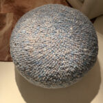 Ronde, blauw gemêleerde Perletta pouffe van gevlochten wol op een lichte vloer.