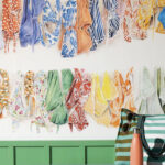 Kleurrijk Pierre Frey La Baignade-behang met illustraties van badkleding aan een wand boven een groene bank met strandaccessoires.