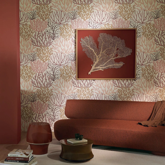 Sfeervolle woonkamer met Pierre Frey behang Acropora FP009001 in koraalmotief, warme rood- en aardetinten en een modern kunstwerk aan de muur.