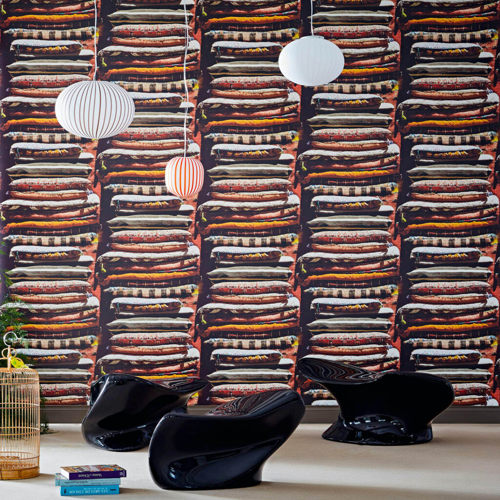 Moderne interieursetting met Pierre Frey-behang FP624001 Le Grand Bazar, waarop kleurrijke stapels kussens zijn afgebeeld.