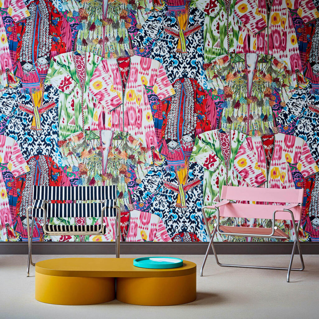 Kleurrijk Pierre Frey-behang FP626001 Manisa met een patroon van traditionele kledingstukken, achter moderne designstoelen en een ovale salontafel.