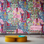 Kleurrijk Pierre Frey-behang FP626001 Manisa met een patroon van traditionele kledingstukken, achter moderne designstoelen en een ovale salontafel.