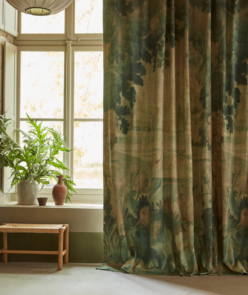 Gordijn van Pierre Frey-stof “Au Bord du Lac” met een natuurtafereel, hangend in een rustiek interieur met een houten bankje en aardewerken vazen.