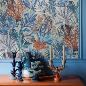 Detail van een woonkamer met het Pierre Frey behang 'Pleine Mer' in blauw, met oceaanmotieven en bijpassende keramische decorstukken op een oranje tafel.