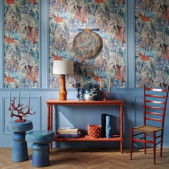 Sfeervolle woonkamer met Pierre Frey behang "Pleine Mer" in blauw en roodtinten, gecombineerd met kleurrijke meubels en maritieme decoratie.