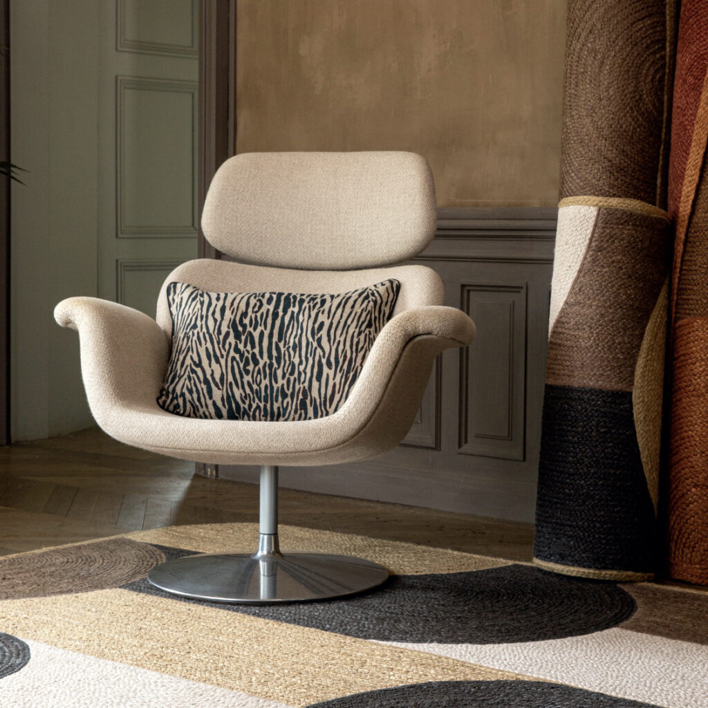 Beige designstoel met metalen voet, voorzien van een sierkussen met dierlijk patroon, in een stijlvol interieur met opgerolde vloerkleden.