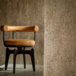 Een designstoel met leren zitting en rugleuning staat naast een wand bekleed met het goudkleurige mozaïekpatroon Tessera2-GXAI-3.