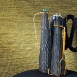 Twee smalle flessen in zwart gevlochten omhulsel uit de VP-711 Dandy serie, staand op een zwarte ondergrond tegen een goudkleurige achtergrond.