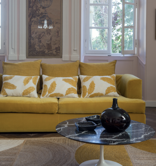 Gele bank met sierkussens, een zwart marmeren salontafel en het ronde vloerkleed Rocky van Elitis in een sfeervolle woonkamer.