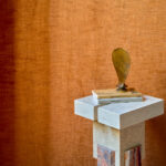 Decoratief stilleven met boeken en sculptuur op een marmeren zuil, voor een warmoranje Alcove 2 RM 410-70 gordijn.