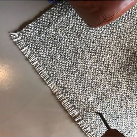 Grijs Perletta Doppio vloerkleed met franjes op een gladde vloer naast een bruine leren stoel.