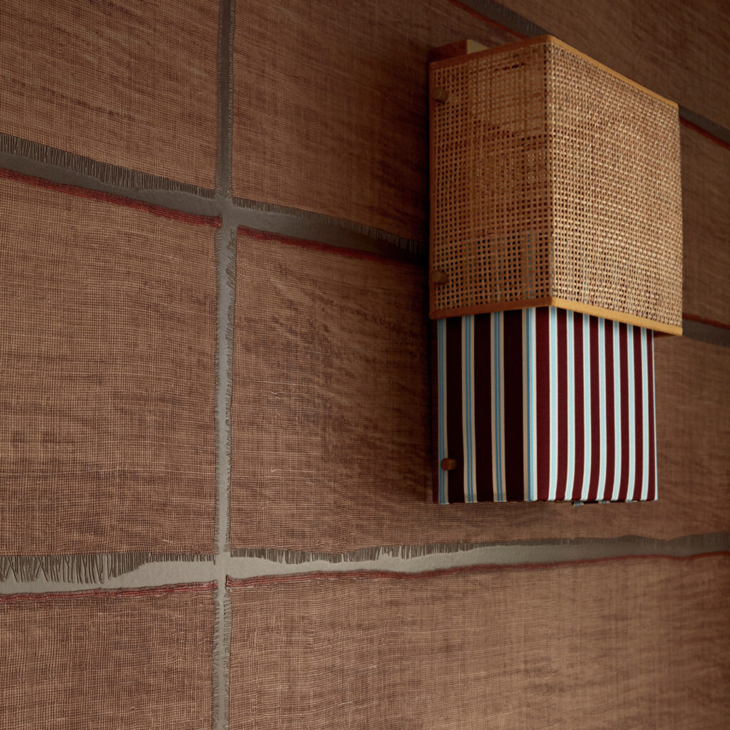 Wandlamp Moshi Moshi van Caravane met gevlochten kap en gestreepte stof tegen een bruinrood textuurbehang.