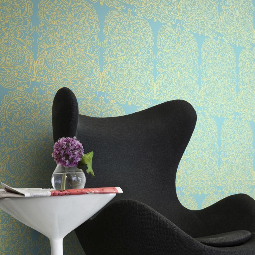 Zwarte designstoel naast een wit bijzettafeltje met vaas en paarse bloemen, tegen Cole & Alpana behang met goudgeel patroon op een blauwe achtergrond.