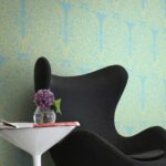 Zwarte designstoel naast een wit bijzettafeltje met vaas en paarse bloemen, tegen Cole & Alpana behang met goudgeel patroon op een blauwe achtergrond.