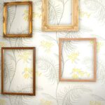 Decoratieve lijsten hangen op een muur met Cole and Son Mimosa-behang met grijsgroene bladeren en gele bloemen.