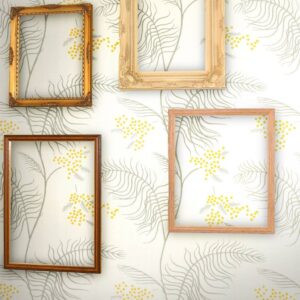 Decoratieve lijsten hangen op een muur met Cole and Son Mimosa-behang met grijsgroene bladeren en gele bloemen.